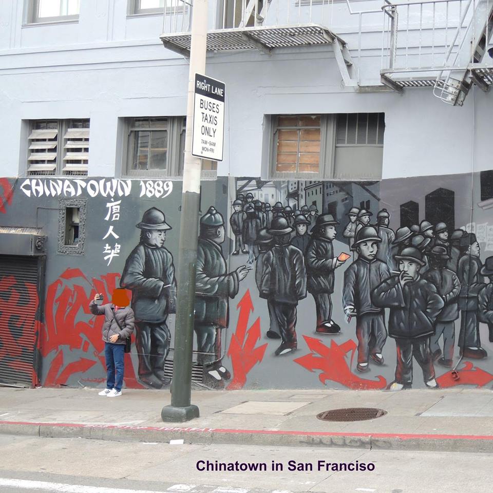 Das Foto zeigt ein großes Wandgrafiti mit grauen Figuren, die Chinesen darstellen sollen (erkennbar an den Hüten), die rauchen und in Gruppen in den Straßen von Chinatown im San Francisco stehen. Rote Pfeile am Boden weisen den Weg. 