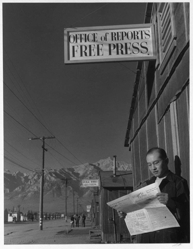 Photo shows editor Roy Takeno reading a copy of the Manzanar Free Press in front of the newspaper office at the Manzanar War Relocation Center; with mountains in the background.